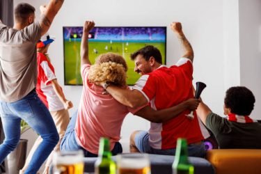 Group of young friends having fun watching football match on TV, drinking beer and cheering; football fans watching game at home celebrating after their team scoring a goal